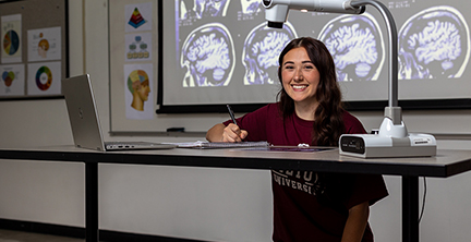 Female student holding a pen and smiling. Surrounding her is a laptop computer, notepad, a light, and in the background is a projection of brain scans.