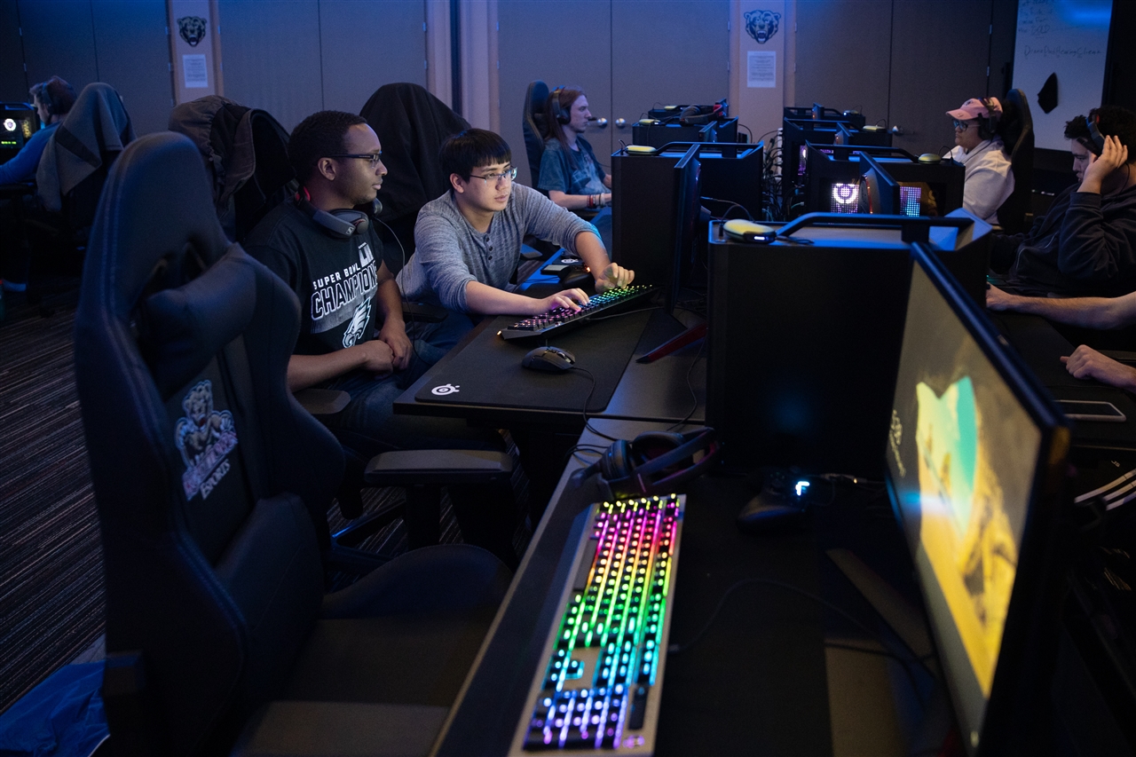 Students playing in the esports room, with one male student assisting another male student in the foreground