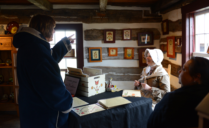 Female wearing traditional clothing, answering visitors' questions from behind a display table 