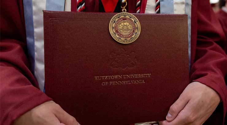 graduate's hands holding Kutztown University diploma case, wearing an academic excellence medal.