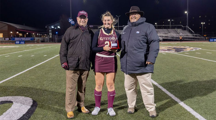 Sara Gatehouse on the field, holding championship scholar plaque. She is accompanied by KU President Hawkinson and PSAC Commissioner Murray