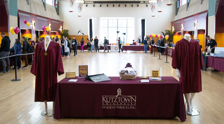 Image of a table with two mannequins dressed in graduation gowns on either side and people walking around gradfest in the background.