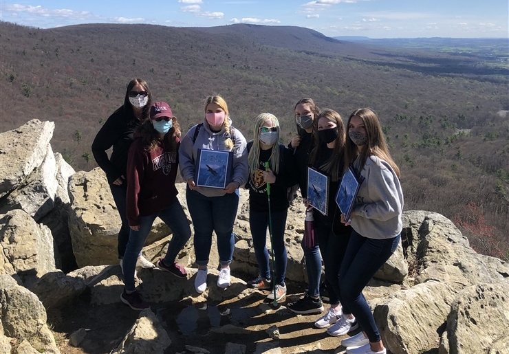 7 people standing on a rock ledge at Hawk Mountain with sky and mountains in the background