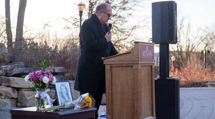 President hawkinson speaking at podium at the Alumni Plaza, with photo and flowers in the foreground