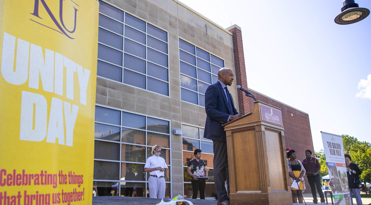 Man standing at a podium outside of a building next to a yellow banner that reads KU Unity Day Celebrating the things that bring us together