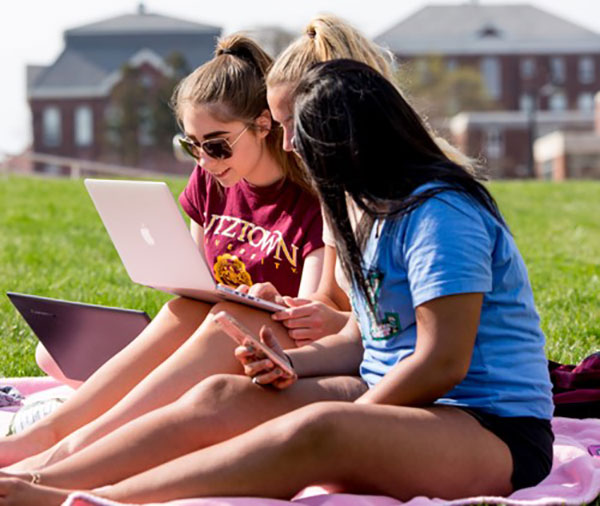 three female students sitting on the DMZ lawn on a sunny warm day laughing and conversing while looking at a laptop screen