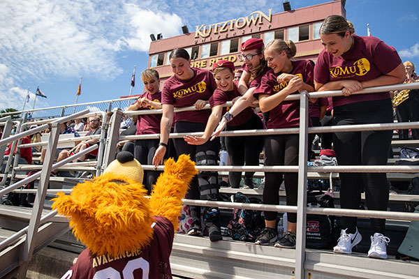 Our KU mascot Avalanche the golden bear reaching up to a group of fans in the stands of the football stadium giving high fives.