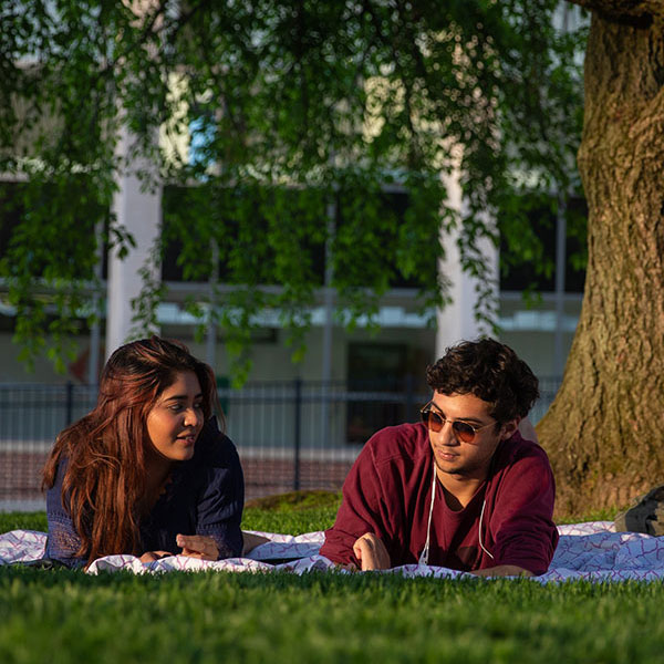 Two students one female and one male laying on a blanket on a lush green lawn conversing. A stately green tree and the front of the library windows in the background.