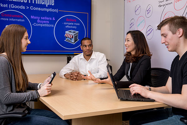 A group of students seated with a professor around a table conversing. One white female looks at a tablet. A black male listens intently hands folded. An Asian professor explains the diagram on the screen behind them. A white male types notes on his laptop.