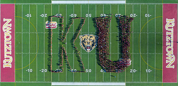 An arial shot of the Andre Read Stadium Filed from end zone to end zone with the golden bear logo on the field in the middle. Student stand in formation to create the letters K and U