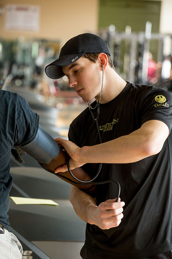 A white male student wearing a black baseball cap and a black KU athletics t-shirt examining an arm of another student in a sports med classroom.