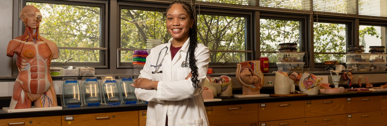 An african american femal student standing arms crossed in her white medical coat in front of various health anatomy figures and supplies