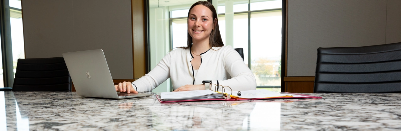 A female sitting at a boardroom table with a laptop and files on the table. She is writing on a piece of paper but looking at the camera and smiling.