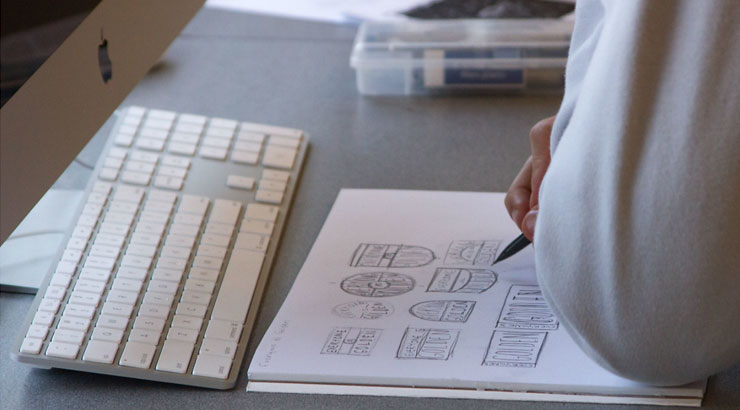 image of a MAC computer and keyboard with a paper sketch book and hands sketching thumbnail designs of logo ideas