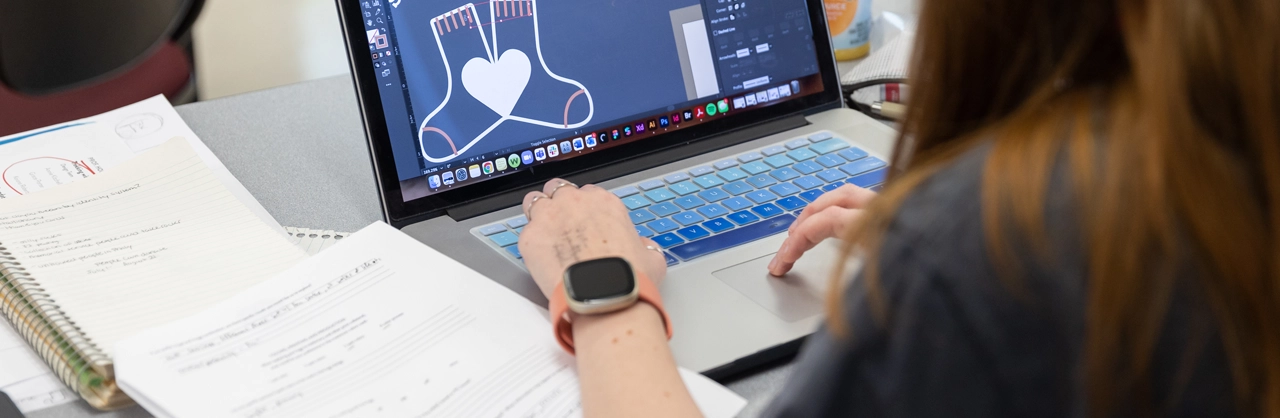 A student sitting at a desk designing a logo for socks on a computer screen and sketches on the table