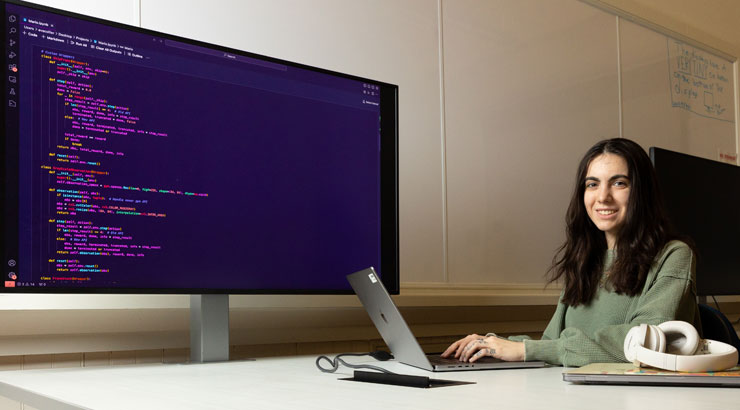 A female computer science student sitting at a desk with a laptop and a large screen with code on it