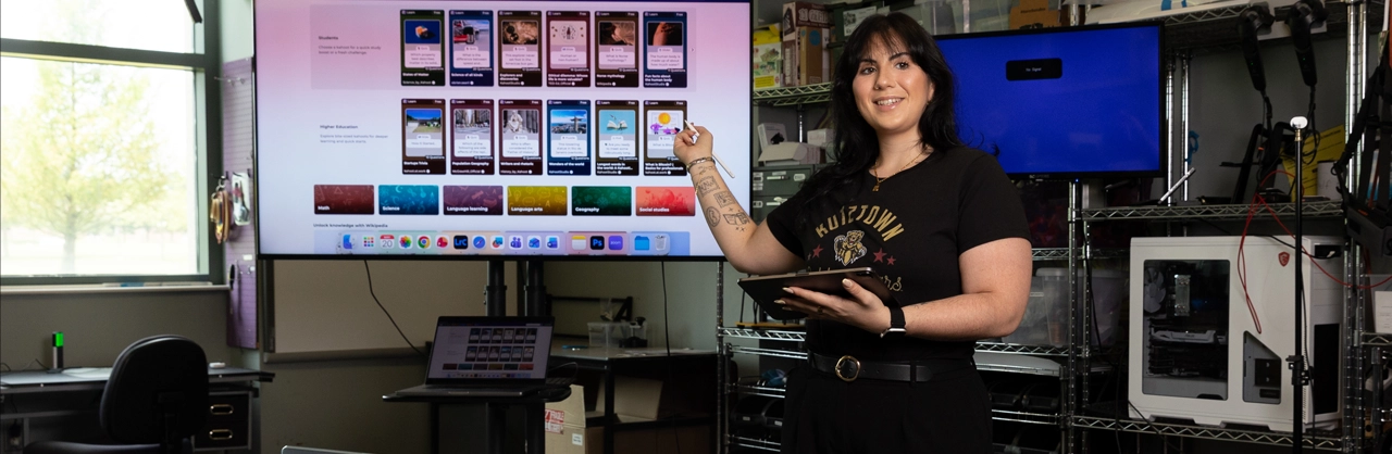 A woman standing holding a tablet while also pointing at a screen behind her instructing a class