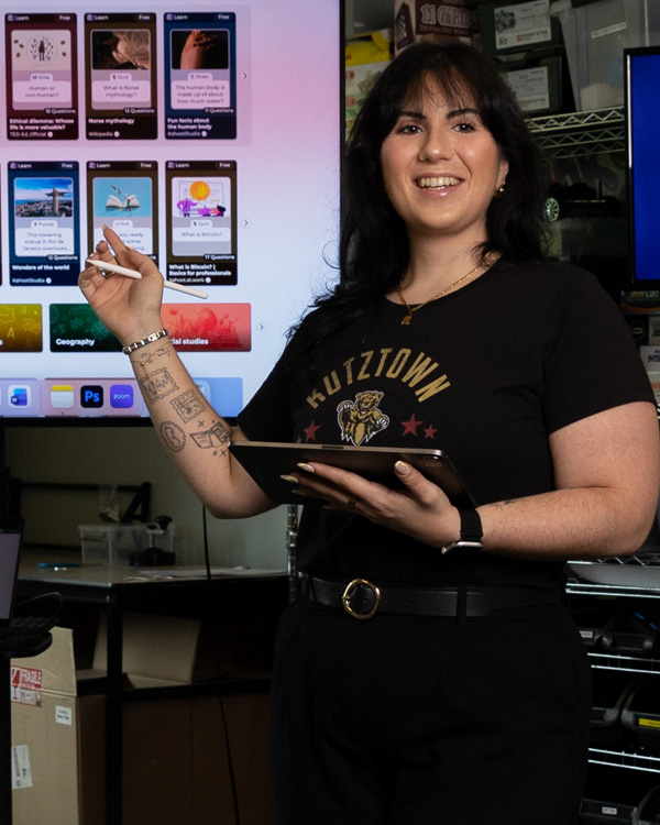 A woman standing holding a tablet while also pointing at a screen behind her instructing a class