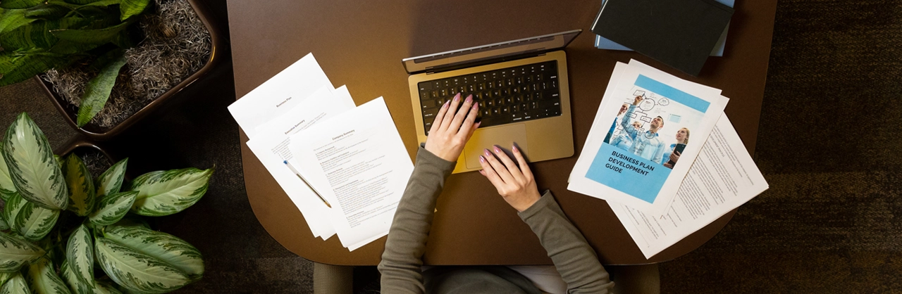 An image from above of hands typing on a laptop with a business plan development guide and other business plan papers on the sides