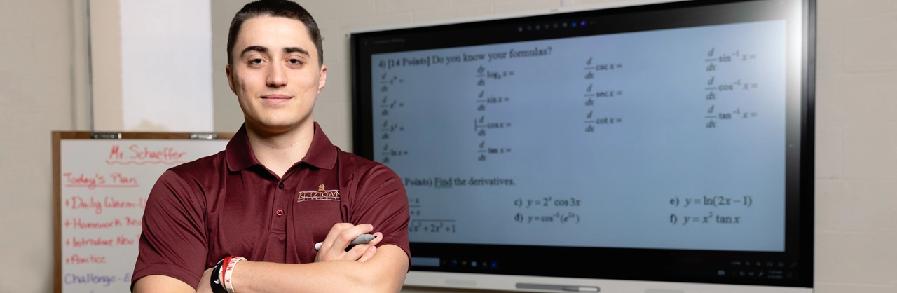 A male student standing posed with his arms crossed in front of a smartboard with math problems on it