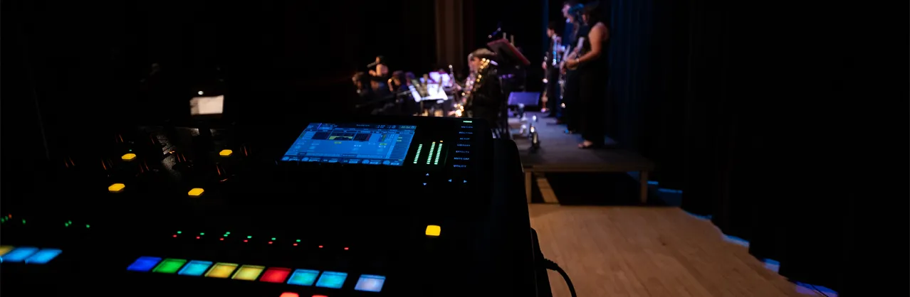 Outline of a male student at a lite up sound board back stage with musicians playing a concert on the stage