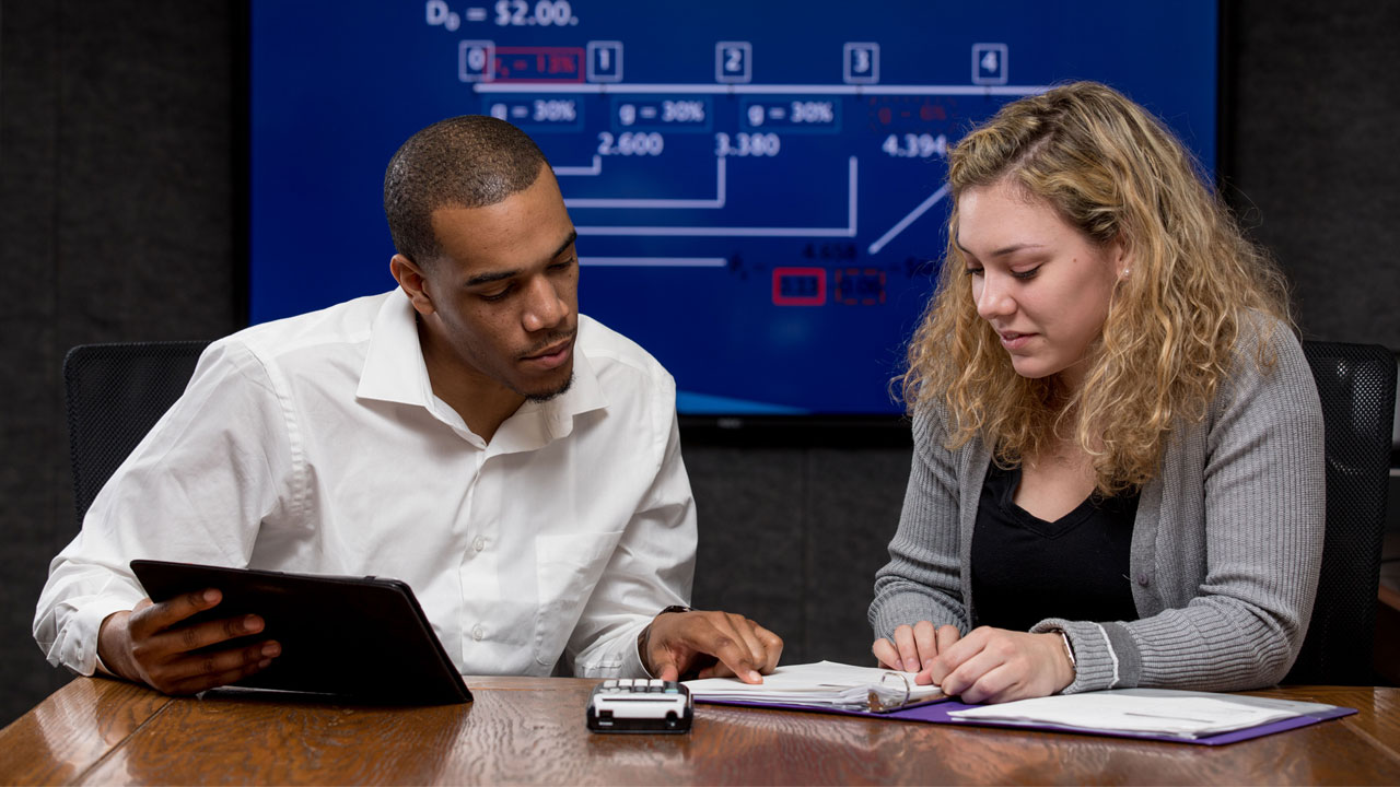 Male student on left, holding tablet and pointing at a book that sits in front of a female student, who is looking at the book.  In the background is a screen that shows diagrams and business wording.