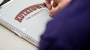 An image of a hand holding a pen over a notebook with the words kutztown university printed on it